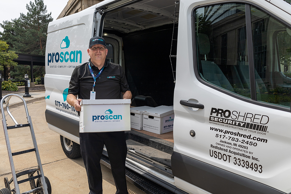 A Proscan employee, dressed in a sleek black uniform and matching cap, confidently holds a box of documents. Behind him, the Proscan van stands ready with its door open, revealing neatly stacked file boxes inside.